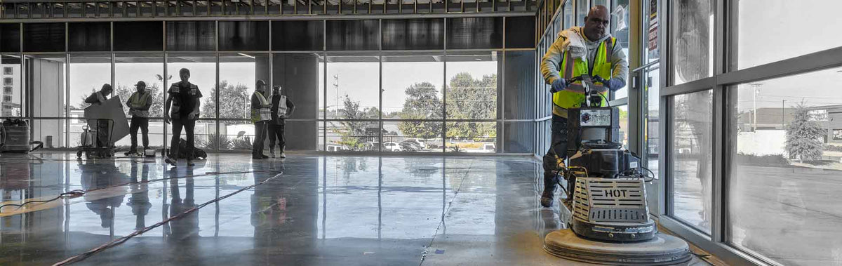 A worker in a high-visibility vest operates a floor burnisher on a polished concrete surface inside a large, open commercial space with large windows, while other workers stand in the background.