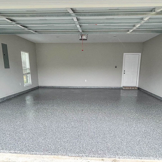 A large, empty garage interior with a freshly installed speckled gray epoxy floor,  a white door on the right, and a window on the left.