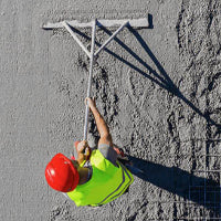 A construction worker in a red hard hat and bright yellow safety vest uses a large metal screed board to smooth wet concrete on a construction site.