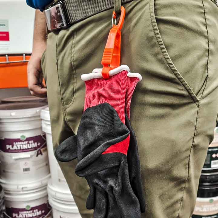 A close-up view of a hi-vis orange glove clip attached to a worker's belt loop, holding a pair of black work gloves against khaki pants.